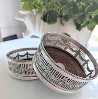 Pair of silver plated wine coasters with pierced decorative sides and wooden inserts on a white surface with a plant and elephant ornament in background.
