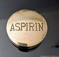 Top view of the brass paperweight showing 'ASPIRIN' embossed text, reflective surface with minor wear.