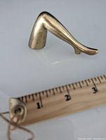 Side view of the vintage brass bottle opener shaped like a female leg, displayed next to a ruler showing length around 3 inches.