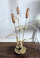 Full view of the large metal sculpture showing three cattails on tall stems with curved leaves and round base leaves in gold and brown tones, displayed indoors against a wall.