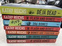 Photo showing spines of seven Kathy Reichs hardcover books stacked horizontally. Titles visible include 'Déjà Dead', 'Flash and Bones' (two copies), '206 Bones', 'Bones Are Forever', 'Devil Bones', and 'Spider Bones'.