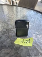 Photo showing a black rectangular Zippo lighter standing upright on a glass table. The lighter features the white Zippo logo printed vertically on its front side.