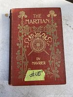 Front cover of antique book The Martian by George du Maurier featuring red cloth binding with gold embossed floral and compass design, and title and author name.