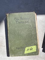 Front cover of an antique book titled The Son of Tarzan with author name Edgar Rice Burroughs in black text on green textured cloth cover. Yellow sticker with lot number
