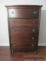 Front view of antique mahogany highboy showing five drawers with round metal handles, standing on rounded legs against a white wall and wood floor.