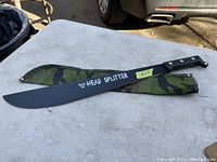Photo of the black machete with 'HEAD SPLITTER' printed on the blade, lying next to its green and black camouflage sheath on an outdoor surface.