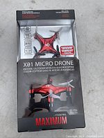 Front view of the X01 Micro Drone in box showing drone with red and black color scheme and propellers. Box front with branding and feature highlights.