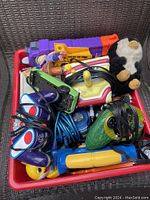 Red plastic bin filled with assorted kids toys including electric handheld toys with cords, colorful plastic toys, and a plush toy.