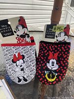 Two oversized Minnie Mouse themed oven mitts, one white with text design and one black with polka dots and Minnie print, shown outdoors with tags.