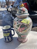 Photo showing a hand painted urn with lid featuring floral and bird designs and a blue Wedgwood jasperware pitcher with white relief designs.