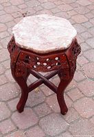 Side angle showing the full carved rosewood plant stand and its octagonal pink marble top resting on the carved base. The stand is set on a paved surface.