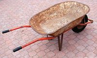 Side view of the metal wheelbarrow showing rusted basin and red handles with black grips on a paved surface.