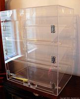 Front and side view of clear acrylic display case showing metal hinges and three slanted shelves inside, on black surface near window.