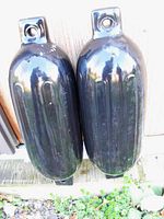 Photo showing two black cylindrical boat bumpers with flat mounting tabs and holes for attachment, placed side by side against wooden and outdoor background.