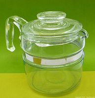 Front view of the coffee pot on a green and yellow background showing clear glass pot with handle and lid.