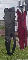Two sleeveless wetsuits hanging outdoors on a line: one black, one red with black and beige accents.