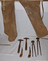 Leather chaps hanging with 6 farrier tools laid out beneath them on a white cloth background.