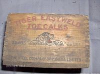 Top view of wooden box showing TIGER Easy Weld Toe Calks advertising with tiger head graphic and weight markings, weathered and faded paint.
