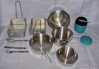 Photo showing full assortment of aluminum camping pans, soup cup with utensils including spoon and fork, and compact cooking utensils arranged on white fabric surface.