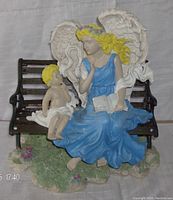 Front view of ceramic angel figurine sitting on a black metal bench with a small child, colorful details including blue dress, yellow hair, white wings, green grass base.