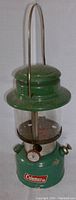 Front view of vintage green Coleman liquid fuel lantern with metal handle, glass globe, and red Coleman logo on the base.