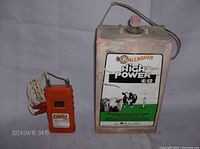Photo showing Gallagher High Power IE12 electric fence charger with metal casing and label with cow image. Adjacent is the orange DVM2 digital electric fence voltmeter with attached wire and clips.