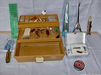 Full view of brown plastic fishing tackle box with two-tiered compartments showcasing various lures, bobber, hooks, and other fishing accessories alongside metal fish holders, scale and line spool.