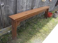 Long solid wooden garden bench made of pressure treated pine, positioned outdoors against a wooden fence on grass.