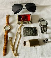 Photo showing men's jewelry pieces including two wristwatches, a pair of black aviator sunglasses, a gold-tone chain necklace, two rings, decorative metal trinket box with red interior, and a brass-colored matchbox-style box.