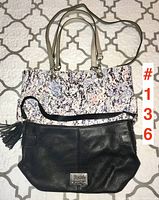 Two Calvin Klein handbags placed on a white and gray patterned bedspread; one black with tassel, one patterned with beige handles.