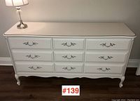 Front view of white French Provincial style dresser with 8 drawers and metal handles, placed against wall with lamp on top