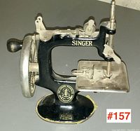 Front angled view of the miniature Singer sewing machine showing the black cast metal base, the hand crank flywheel, and sewing platform with needle and adjustment screws.