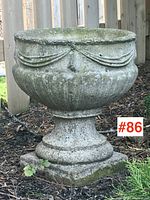Side view of vintage cement garden planter urn outside by a wooden fence, showing draped swag decoration and weathered cement surface