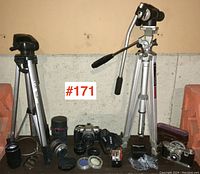 Two tripods, assorted camera lenses, a Pentax MZ-7 camera, and camera accessories arranged on the floor with a brick wall background.