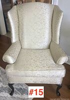 Front view of a vintage wing-back armchair with cream upholstery and subtle gold floral pattern, showing curved arms and padded seat cushion with dark wooden cabriole legs.