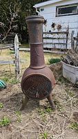 Full view of the upright metal chimenea outdoors with a rusted, brown surface, standing on three legs with mesh door closed.
