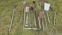 Overview of 11 vintage gardening tools laid out on grass, showing shovels, pitchforks, hoes, and sticks with wooden and metal parts.