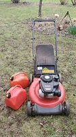Front view of Yard Machines lawn mower with red body, black grass catcher bag, and black handlebar, positioned on grassy ground with two red fuel containers on the left side.