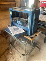 Blue Mastercraft 12.5" thickness planer sitting on a metal stand with a wooden board on the stand's top. Part of the stand's lower shelf is visible with debris on the ground around it.