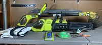 Full view of Ryobi cordless chainsaw, weed snipper, carrying bags, and accessories on table.