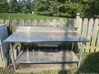 Full front view of the stainless steel commercial kitchen counter showing countertop, drawer, and bottom shelf against wooden fence.