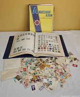 Photo showing two Ambassador brand stamp albums and a spread of loose used stamps from various countries scattered on a white surface.