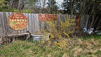 Three vintage metal advertising signs mounted on a fence outdoors among foliage. Rust and weathering evident.