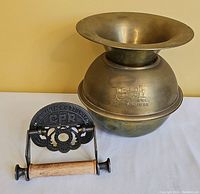 Brass spittoon and cast iron toilet paper holder together on a neutral background.