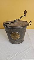 Front angled view of the vintage metal bread maker showing the lid with wooden handle crank and the metal body with a brass plaque.