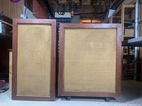 Front view of two vintage wooden speakers, showing fabric covers and wooden frames, one taller and slimmer, the other wider.