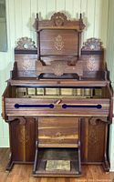 Frontal view of the antique pump organ showing carved wood details, decorative floral inlay, and the front panel partially open with visible internal storage compartments.