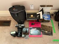 Overview photo showing the Olympus camera with Nikon lens attached, flash unit, Lowepro carrying bag, original boxes, manuals, and photography book arranged on carpet.