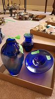 Three cobalt blue glass items including a large pitcher, a small vase, and a dish arranged in a cardboard box on carpeted floor.