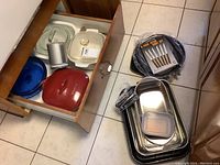 Kitchen floor showing stainless steel roasting/serving trays, cutlery set in box, and a small plastic food container on top